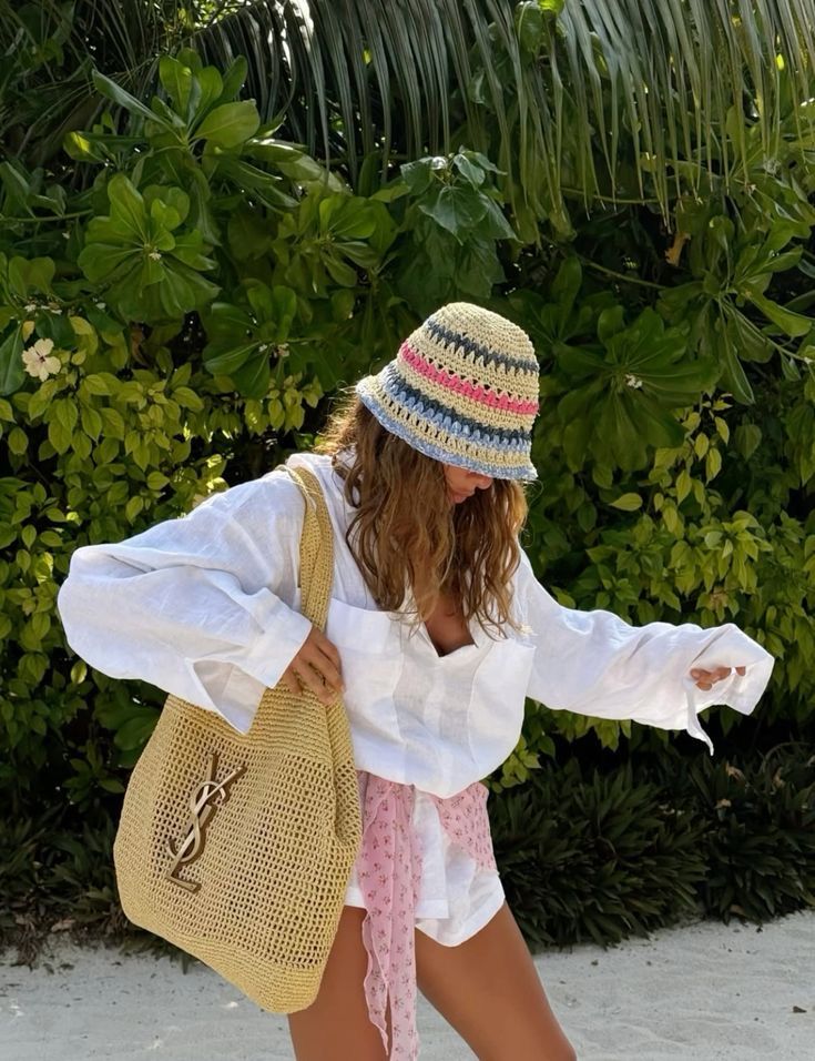 Woman in a colorful hat and beachwear holding a stylish woven bag near lush greenery. Summer fashion, relaxed vacation vibe.