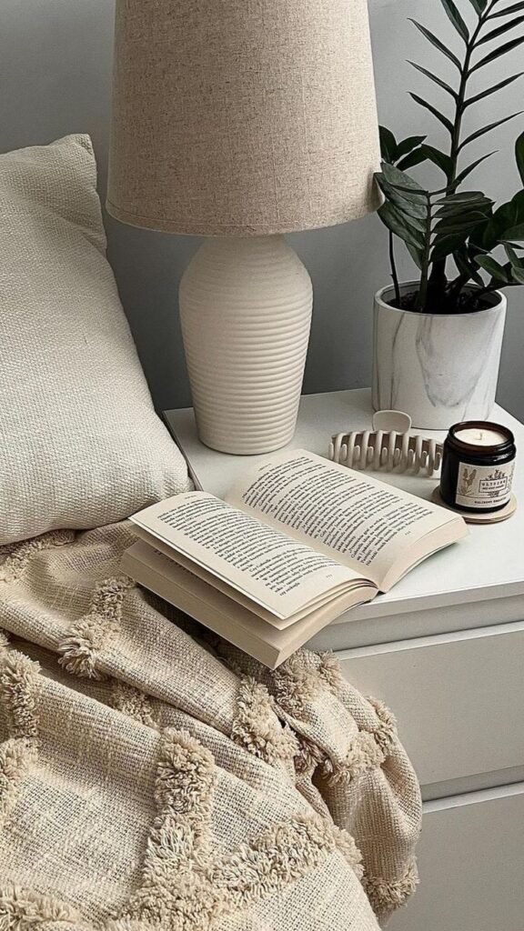 Cozy bedroom scene with open book, beige lamp, candle, plant, and textured blanket on a white bedside table.