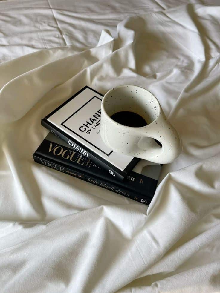Ceramic mug of coffee on stacked fashion books on white bedsheet, blending style and relaxation.