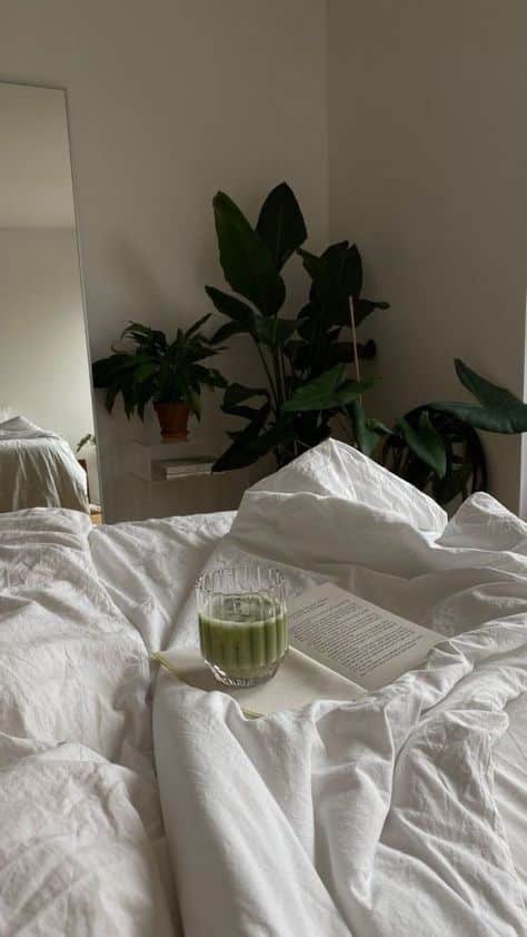 Cozy white bed with a book and matcha latte, surrounded by lush indoor plants for a relaxing morning vibe.