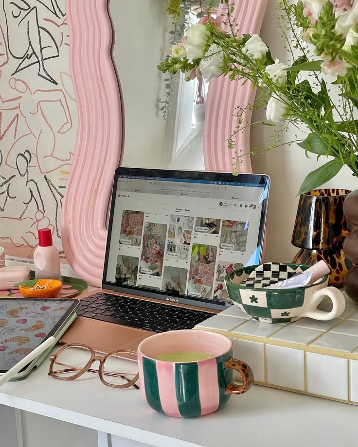 Cozy workspace with laptop, colorful mugs, tablet, and flowers on a desk. Artistic wallpaper creates an inspiring atmosphere.
