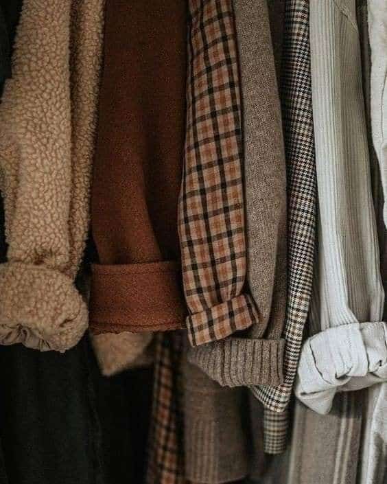 Close-up of cozy fall and winter clothes in warm tones, including plaid, wool, and shearling textures hanging in a closet.