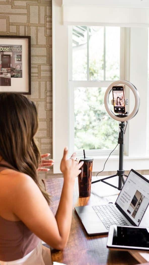 A woman records content using a smartphone on a ring light tripod, with a laptop and tablet on the desk nearby.