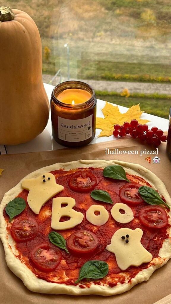 Halloween-themed pizza with cheese ghosts and Boo letters, surrounded by tomatoes and basil leaves. Cozy autumn ambiance.