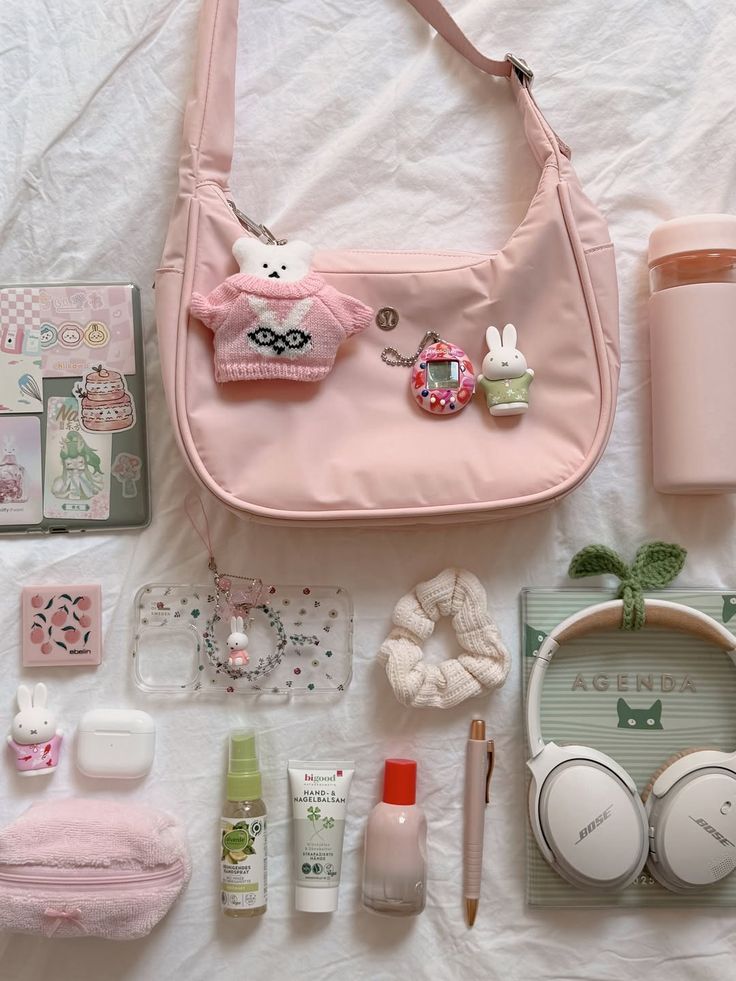 Pink handbag with cute accessories, bunny and bear charms, phone case, scrunchie, headphones, skincare, and planner on white background.