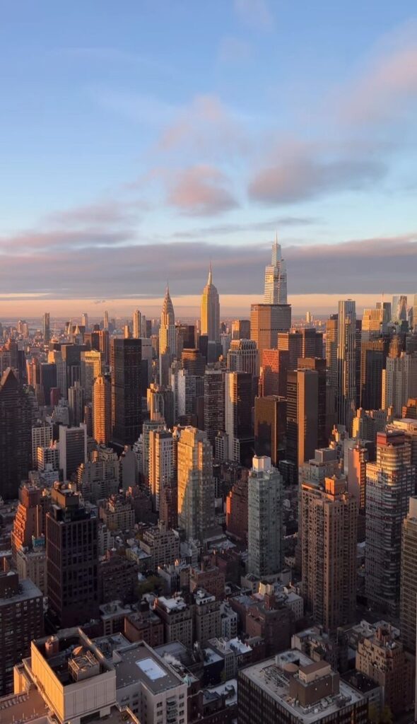 Sunset over a vibrant city skyline with iconic skyscrapers glowing in warm light, under a clear sky with soft clouds.