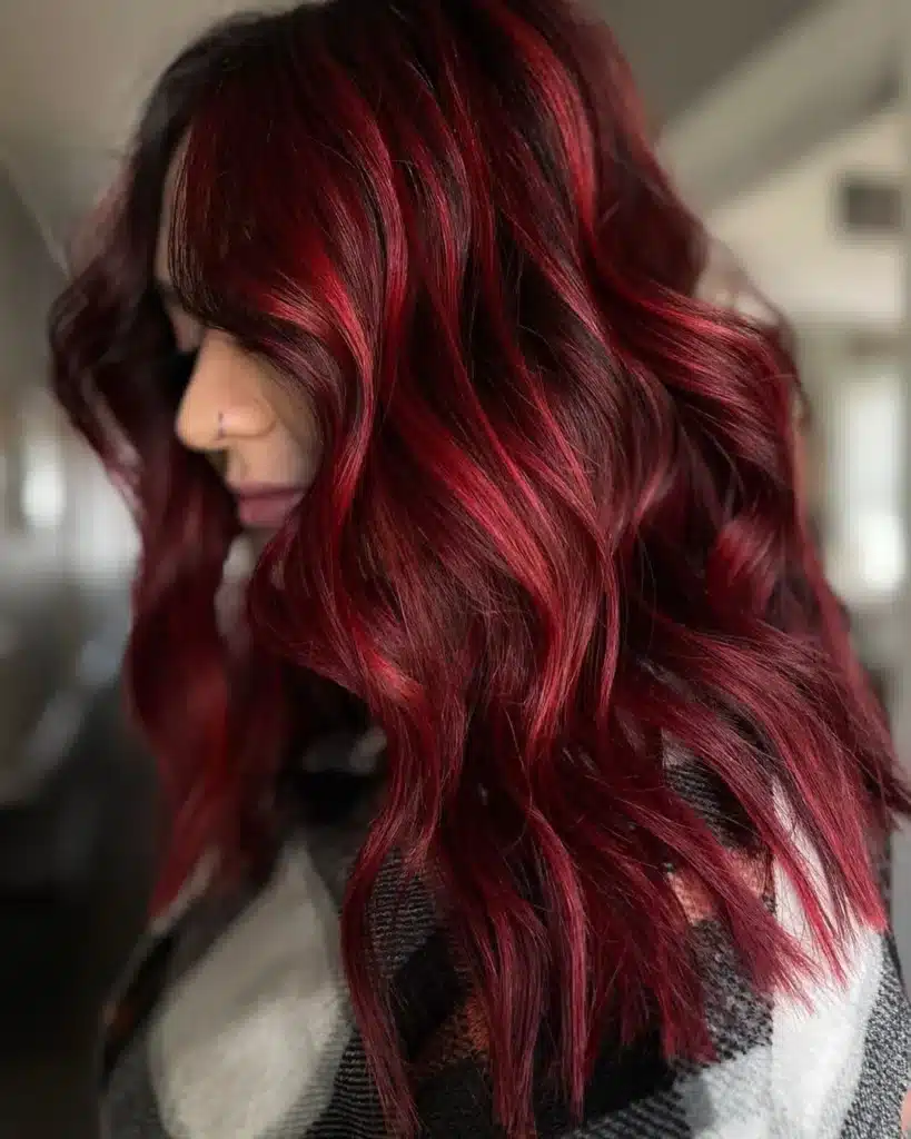 Woman with vibrant red, wavy hair and a nose piercing, wearing a checkered outfit. Hair color stands out with rich, deep hues.