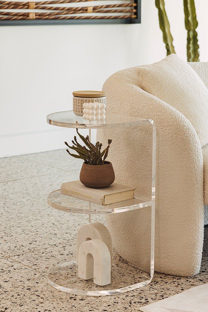 Clear acrylic side table with decorative items and a plant, next to a cozy beige sofa in a modern living room setting.