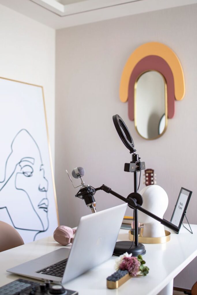 Modern home office with podcast setup including laptop, microphone, and headphones on a white desk with abstract wall art.