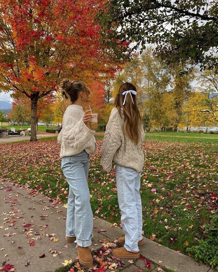 Two people in cozy sweaters enjoy a fall day, surrounded by colorful autumn trees and fallen leaves in a park.