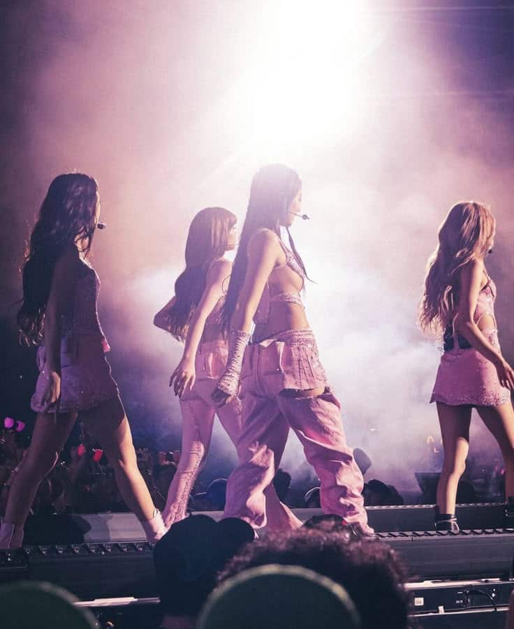Four performers in pink outfits on stage with bright lights and fog, captivating a lively audience.