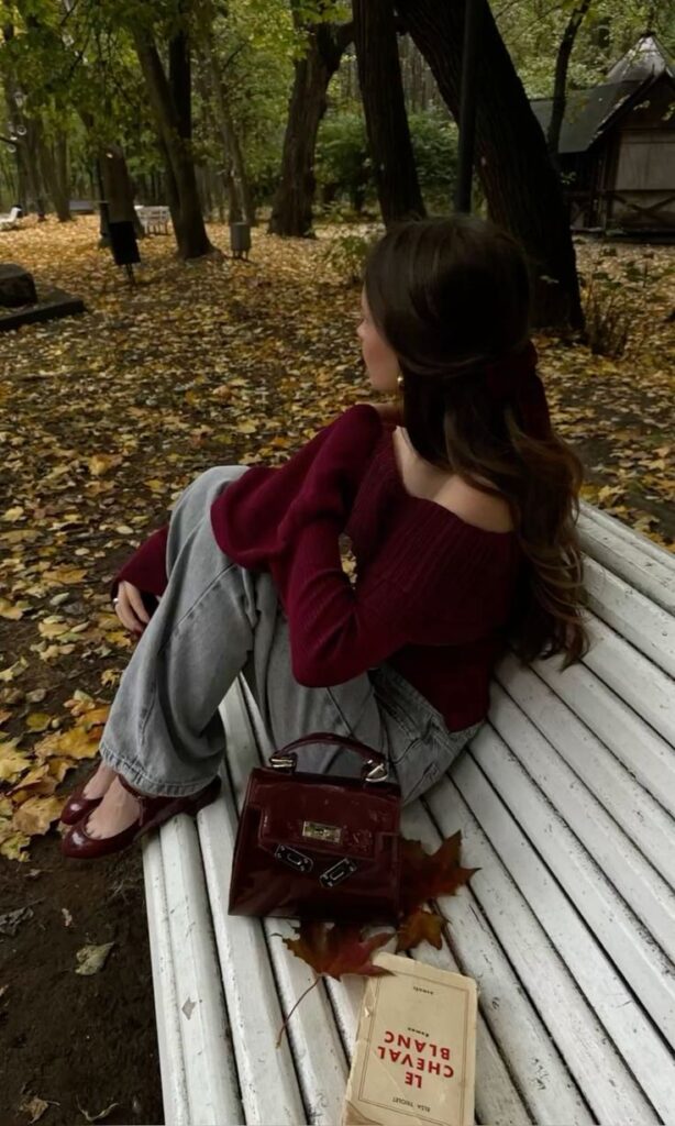 Woman in maroon sweater sitting on a park bench with a book and handbag, surrounded by autumn leaves.