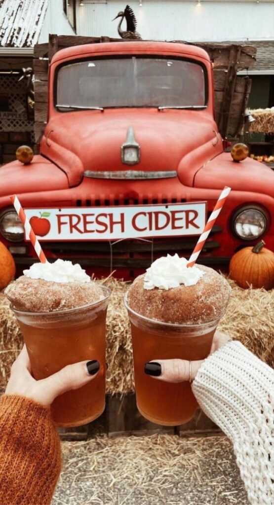 Autumn scene with two apple ciders topped with donuts, held in front of a rustic red truck and Fresh Cider sign.