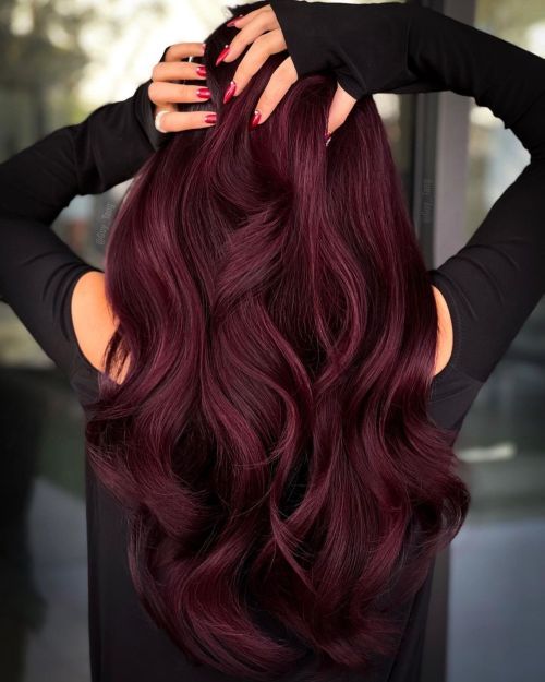 Long wavy burgundy hair cascading over shoulders, nails painted red, hands adjusting strands. Stylish and vibrant hairstyle idea.