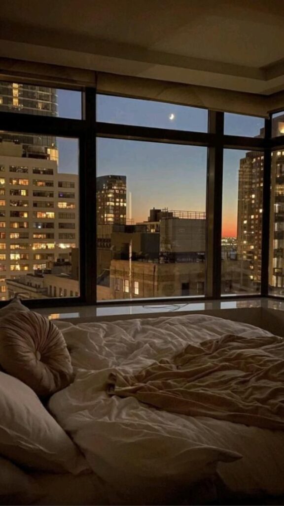 Cozy bedroom with large windows, city skyline view, and a crescent moon at dusk, creating a serene and relaxing atmosphere.
