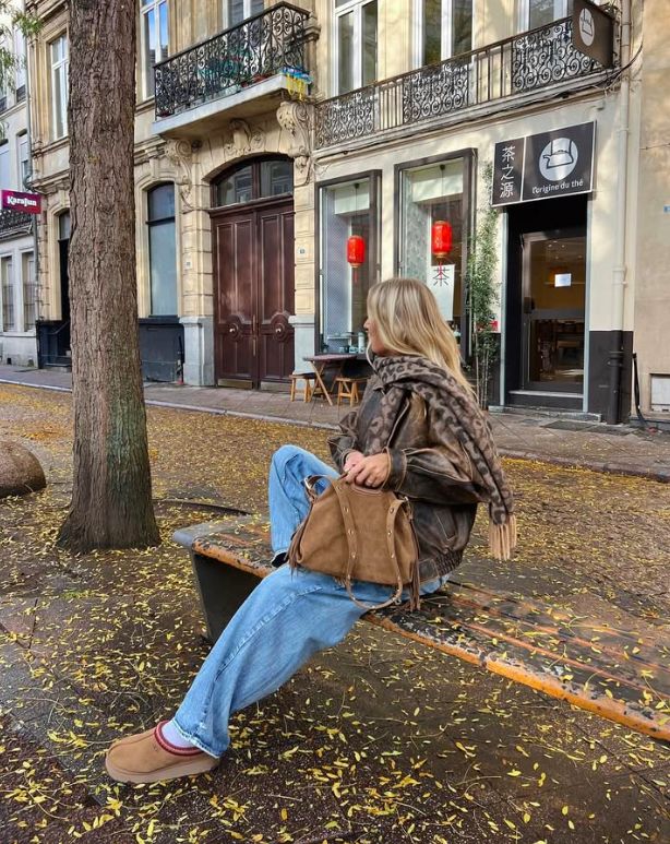 Person sitting on a bench in a leaf-strewn street, wearing casual attire, with a brown bag, in front of a European-style building.