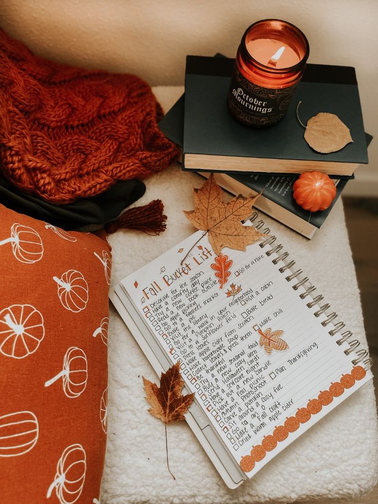 Cozy fall scene with a lit candle, stack of books, and a fall bucket list on a notebook, surrounded by autumn leaves and decor.