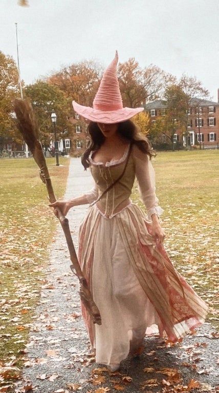 Woman in a vintage witch costume with pink hat and broom walks on a leafy path in autumn.
