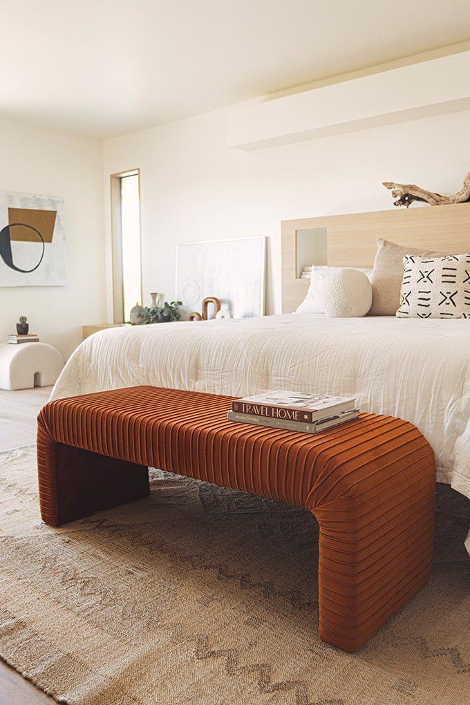 Modern bedroom with a warm-toned bench, cozy bed, abstract art, and soft lighting for a serene, stylish ambiance.