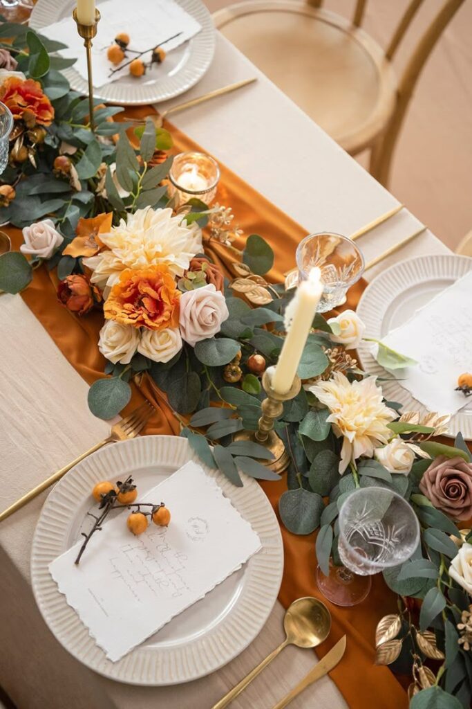 Elegant autumn-themed table setting with candles, flowers, and golden cutlery on an orange table runner, perfect for weddings.
