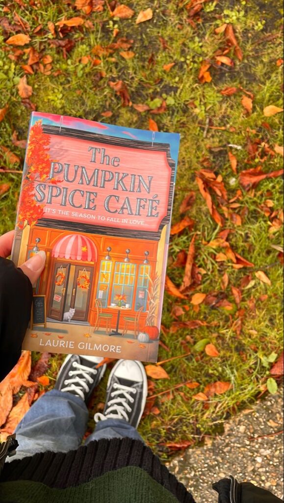 Person outdoors holds The Pumpkin Spice Café book, surrounded by autumn leaves. Cozy fall vibes and literature theme.