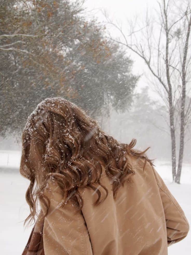 Person with curly hair in a snowy landscape, wearing a tan coat. Trees and snowfall create a serene winter scene.
