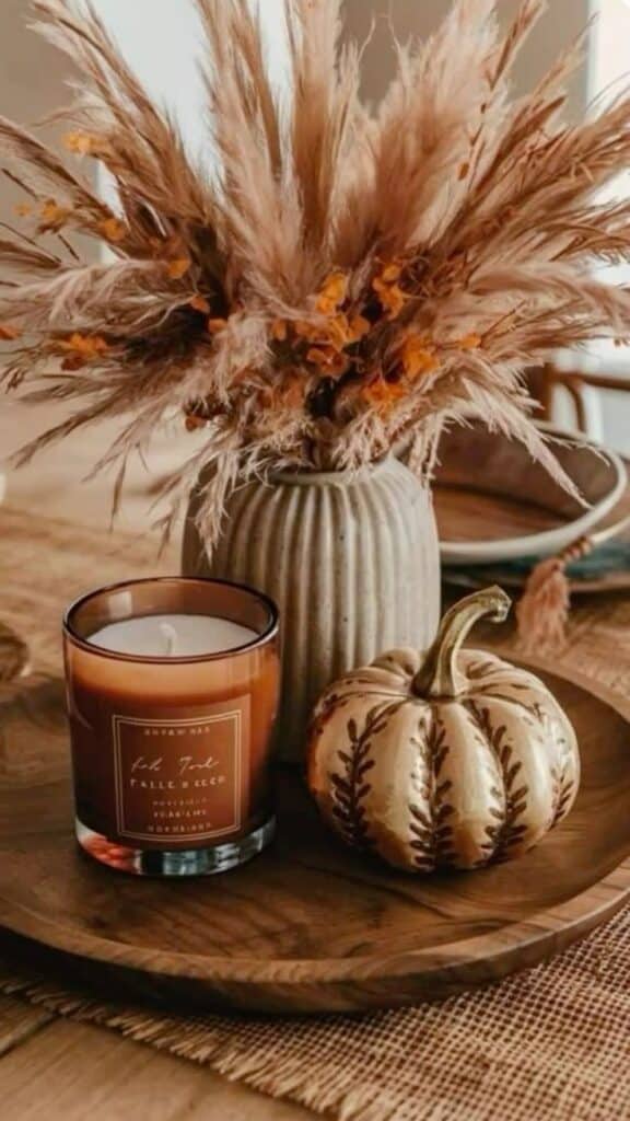 Cozy fall decor with a candle, mini pumpkin, and dried flowers in a vase on a wooden tray, creating a warm autumn ambiance.