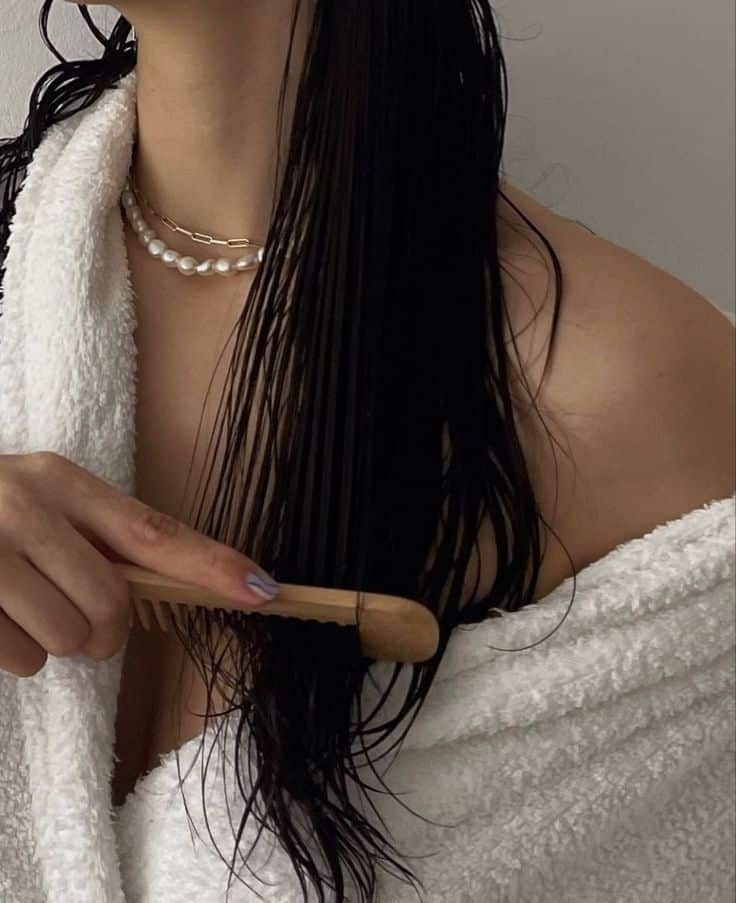 Person combing wet hair with a wooden comb while wearing a towel and layered necklaces.