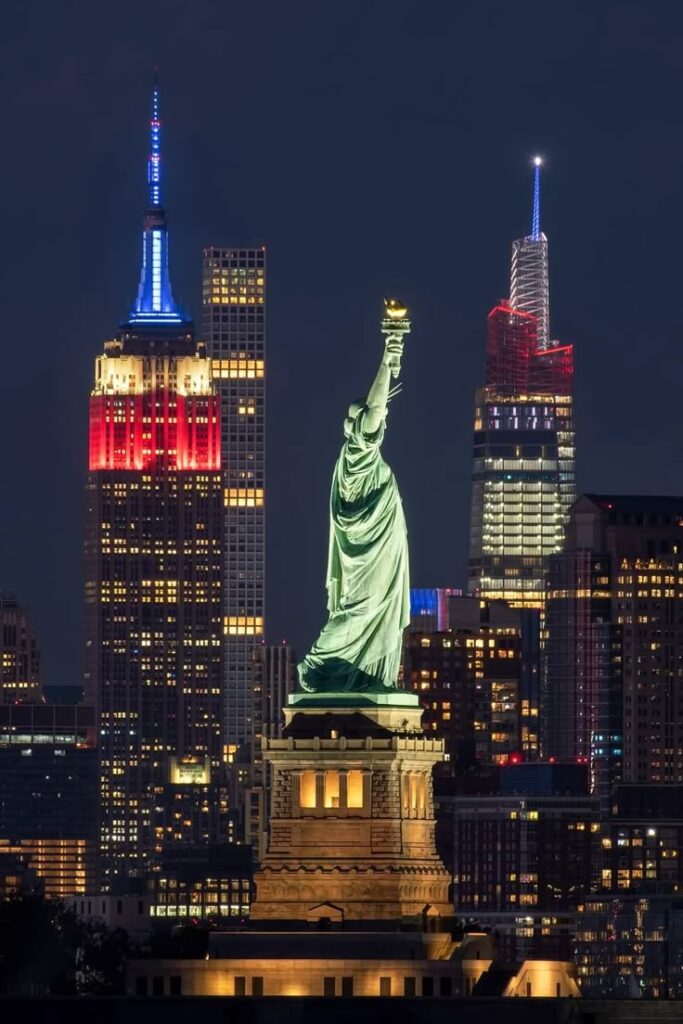 Statue of Liberty illuminated at night with NYC skyline, featuring buildings lit in red, white, and blue. Iconic American scene.