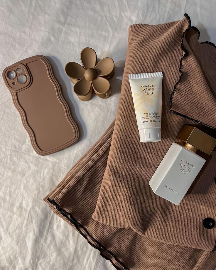 Minimalist flat lay with a beige phone case, daisy accessory, skincare tube, perfume, and ribbed fabric on white background.