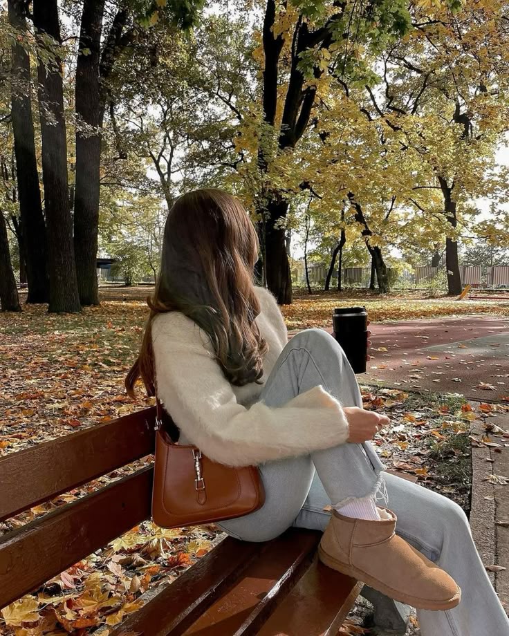 Woman sitting on a park bench in autumn, wearing cozy clothes, surrounded by fallen leaves and trees.