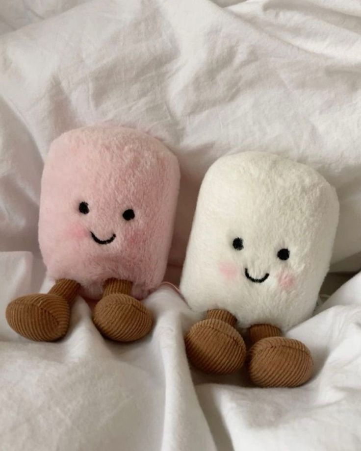 Two cute plush marshmallow toys with smiling faces, sitting on a white bed.