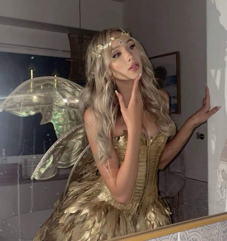 Glamorous woman in golden fairy costume with wings and tiara, posing in front of a mirror at night.