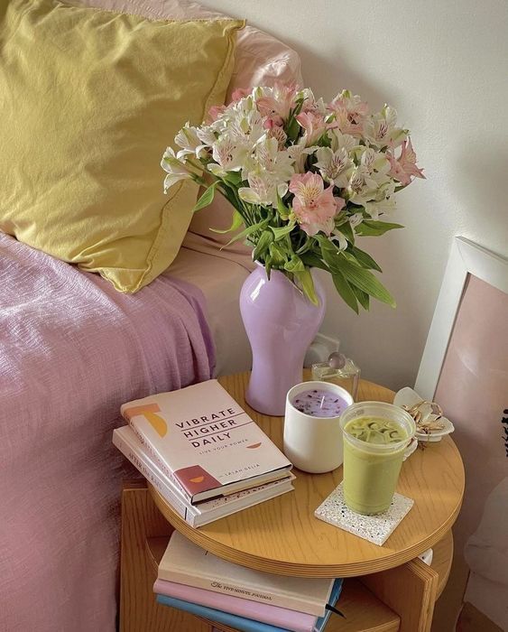 Cozy bedroom scene with pastel pillows, a vase of pink flowers, books, a candle, and iced matcha on a wooden nightstand.