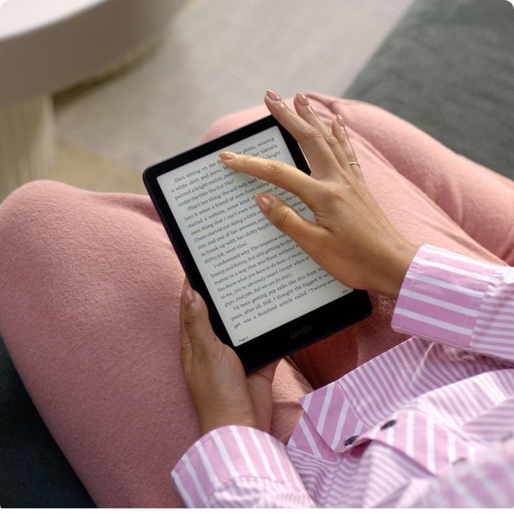 Person reading on a Kindle e-reader while sitting on a couch, wearing pink striped pajamas. Relaxing, digital reading moment.