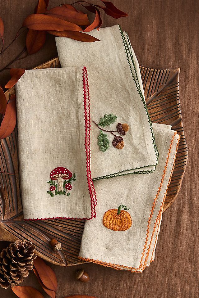 Autumn-themed embroidered napkins with mushroom, acorn, and pumpkin designs, set on a rustic table with leaves and pinecones.