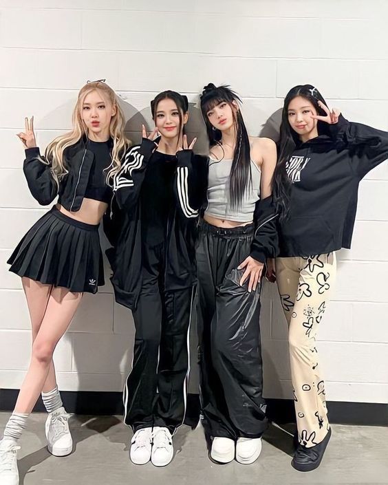 Four stylish women posing in trendy outfits against a white wall, smiling and making peace signs while standing together.