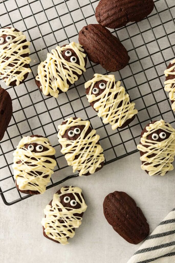 Chocolate madeleines decorated as mummies with white icing and candy eyes on a cooling rack, perfect for Halloween treats.