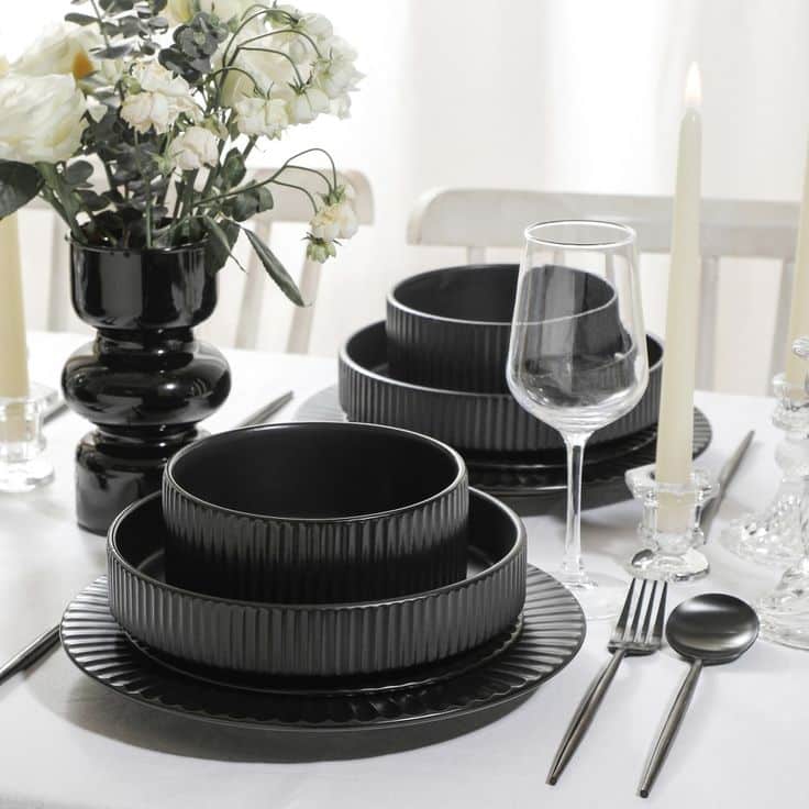Elegant black dinnerware set on a white table with flowers, candles, and glassware for a sophisticated dining setup.