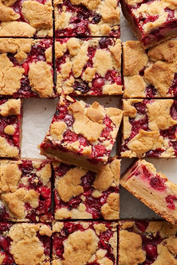 Square cranberry crumb bars with golden crust and vibrant red filling, arranged neatly on parchment paper. Delicious dessert squares.