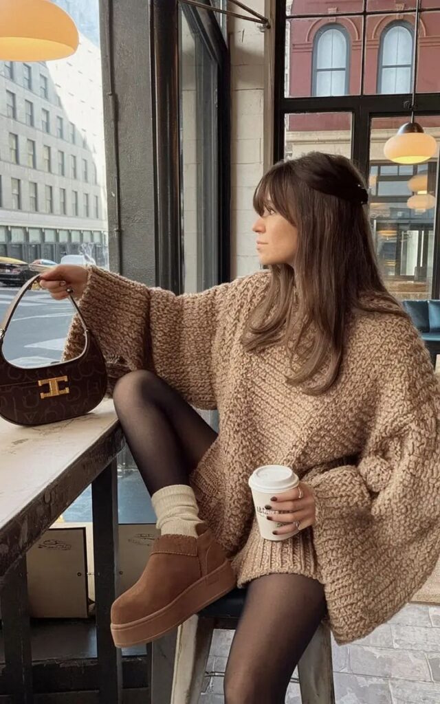 Woman in cozy sweater holding coffee in cafe, wearing brown boots and holding a handbag. Urban winter fashion scene.