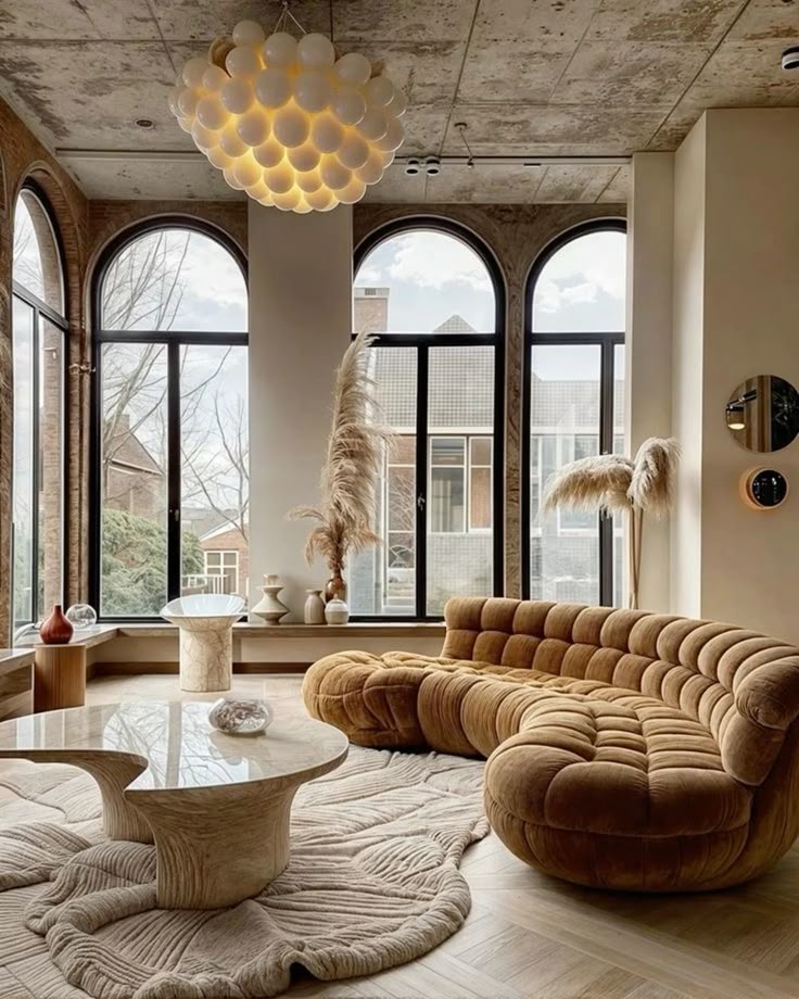 Modern living room with curved brown sofa, elegant chandelier, large windows, and chic decor. Cozy, contemporary interior design.
