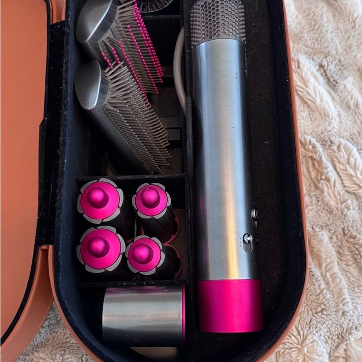 Hair styling tool set with attachments including brush, wands, and dryer in a carry case on soft surface.