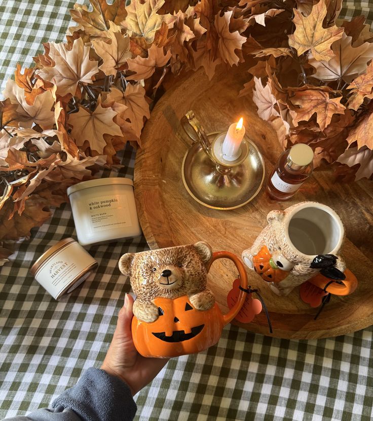 Cozy autumn setup with candles, fall leaves, and cute pumpkin-themed mugs on a checkered tablecloth.