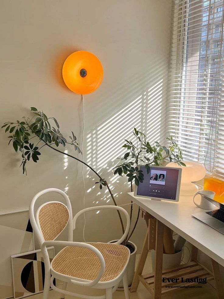 Cozy home office with orange wall lamp, cane chair, plants, and tablet on a desk by a window with blinds letting in sunlight.