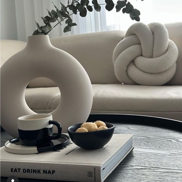Modern living room with cozy decor: circular vase with eucalyptus, knot cushion, coffee on a book, and cookies on a black bowl.
