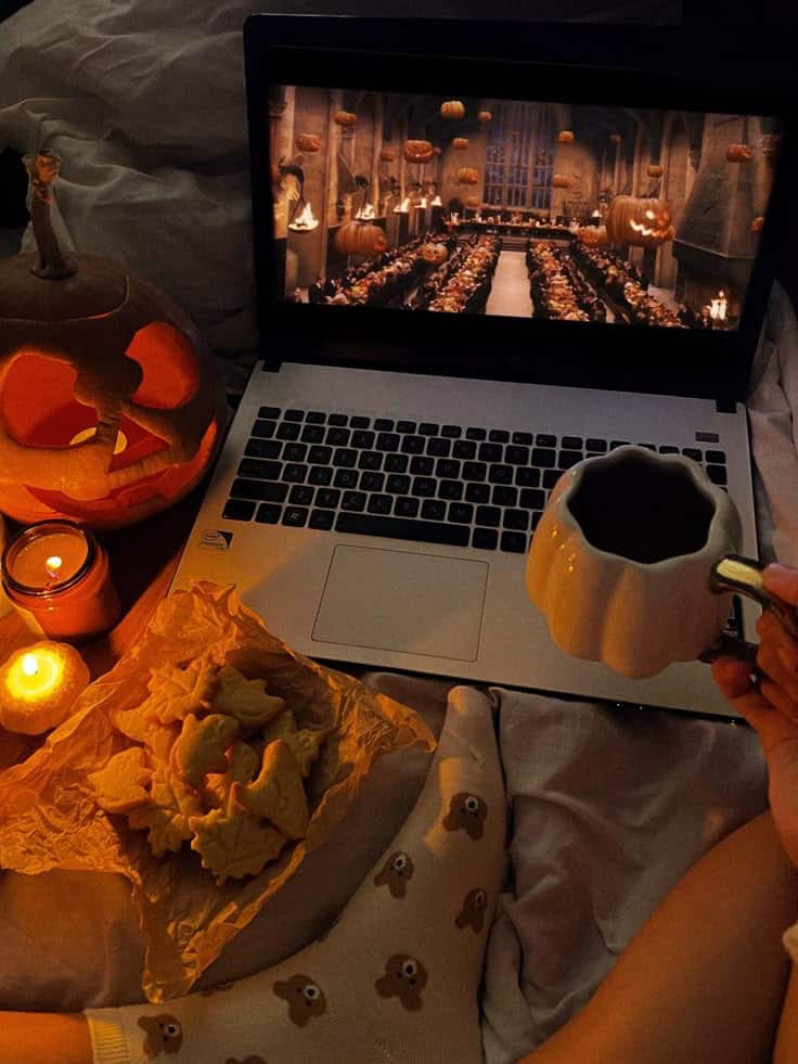 Cozy Halloween movie night with snacks, pumpkin mug, and laptop playing a film. Candles and jack-o'-lantern for ambiance.
