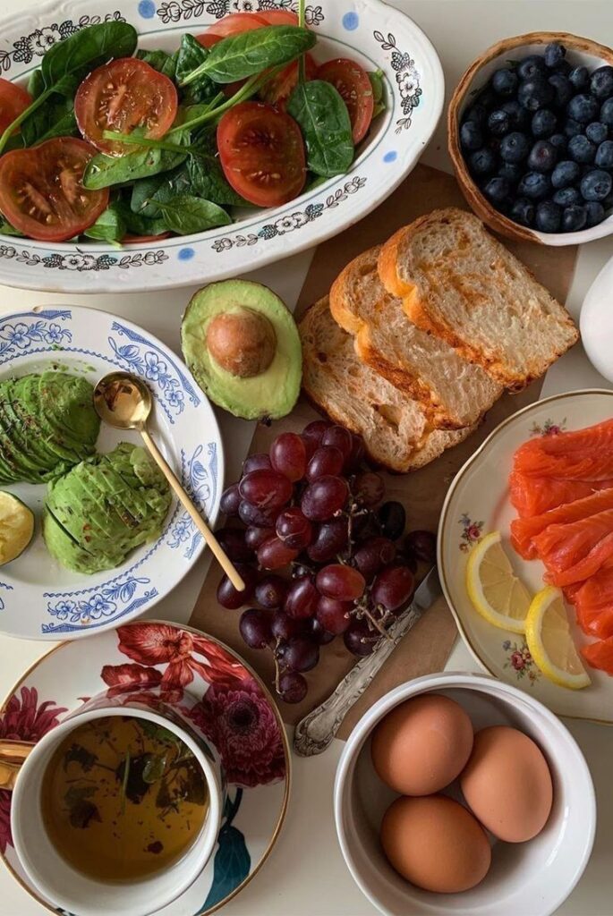Healthy brunch spread with salad, avocado, toast, grapes, berries, eggs, smoked salmon, and tea on a floral-patterned table setting.