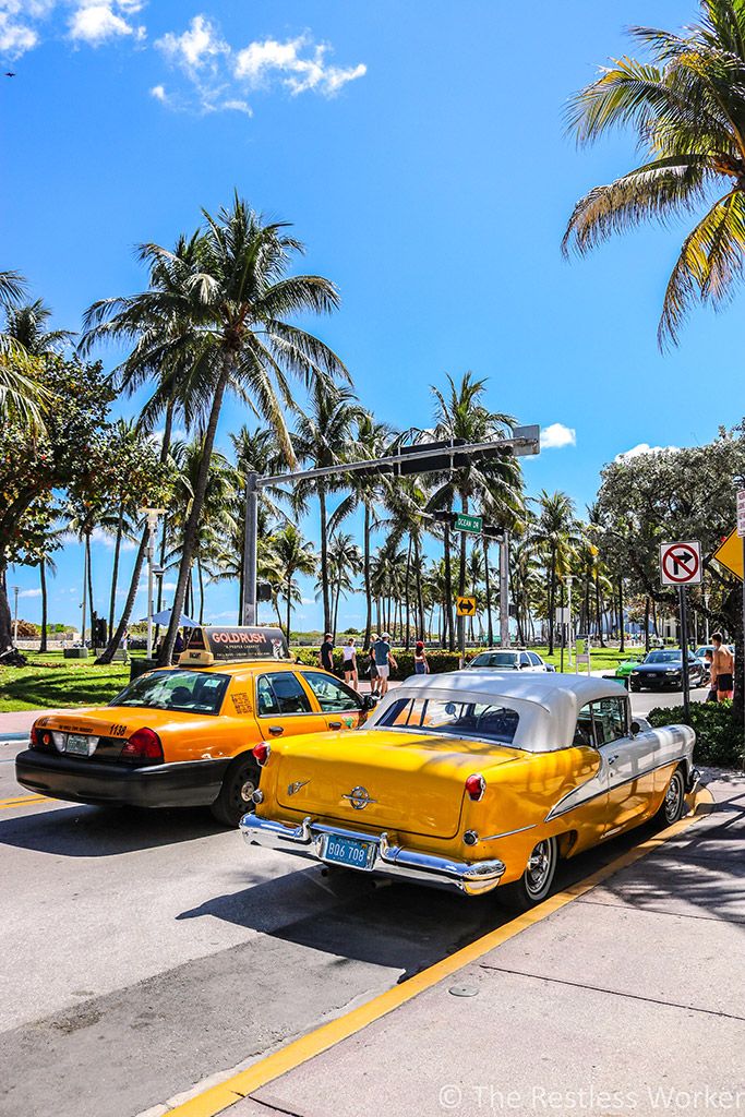 Vintage car and taxi on sunny Ocean Drive, Miami, lined with palm trees under a clear blue sky.
