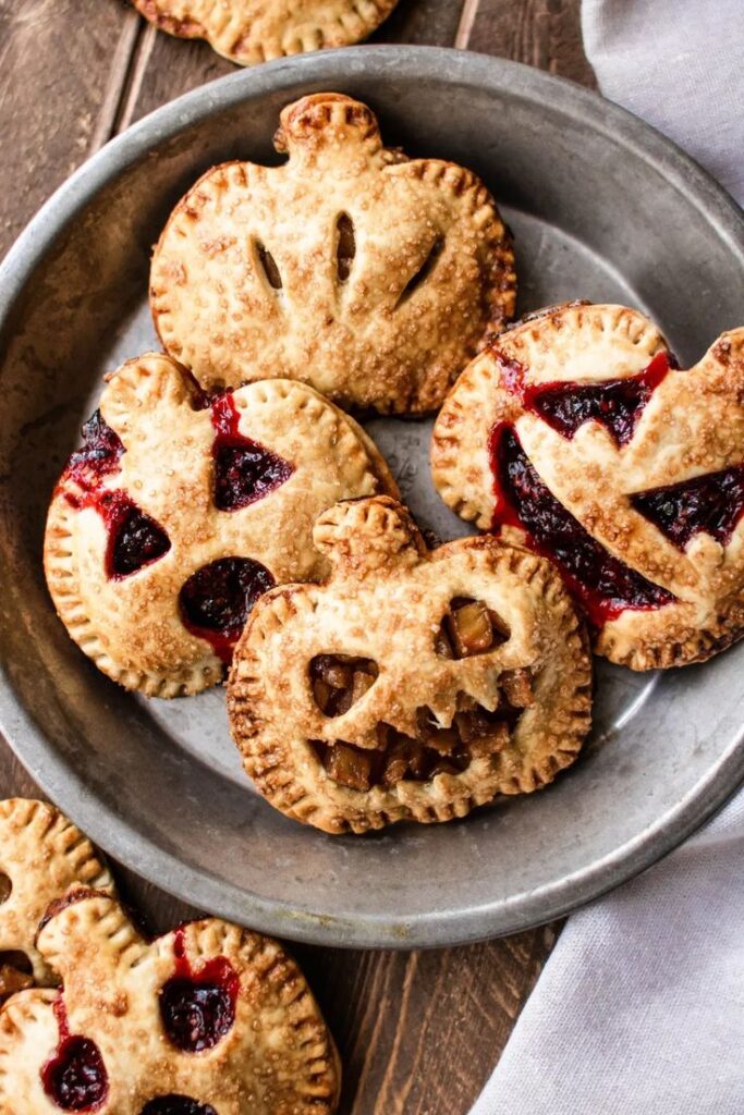Halloween-themed mini pies with jack-o'-lantern faces, filled with jam and fruit, served in a rustic metal dish.